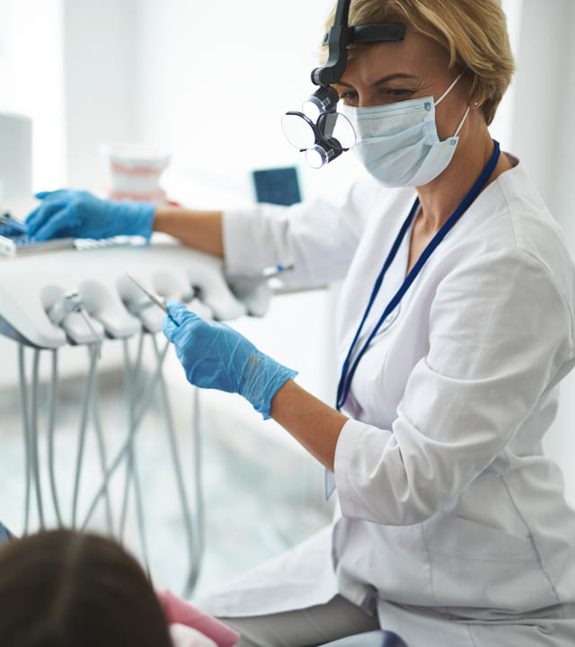 Focus on female specialist standing near chair with girl and looking at her. She is taking tools from holder while wearing uniform and binoculars